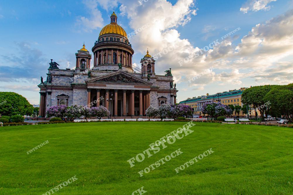 На снимке представлен Исаакиевский собор, расположенный в центре Санкт-Петербурга. Перед ним раскинулась ухоженная зелёная лужайка, обрамлённая цветущими кустарниками. Собор, с его величественным куполом и колоннами, является символом города и одной из его главных достопримечательностей. Летнее время и мягкое вечернее освещение подчёркивают гармонию архитектуры и природы.